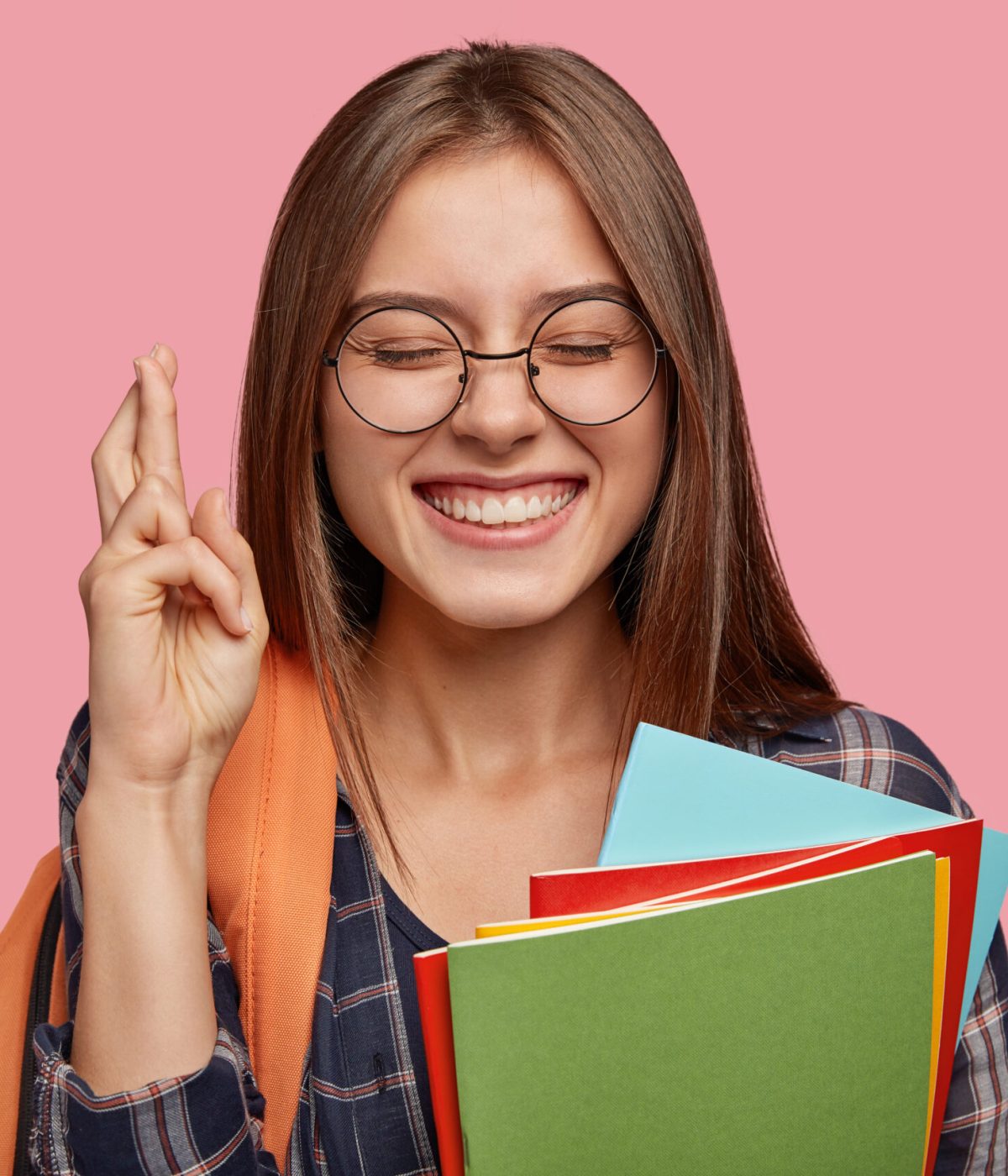 Intense cheerful schoolgirl with broad smile, crosses fingers for good luck, has satisfied expression, keeps eyes closed, carries rucksack, shows toothy smile, isolated over pink background.